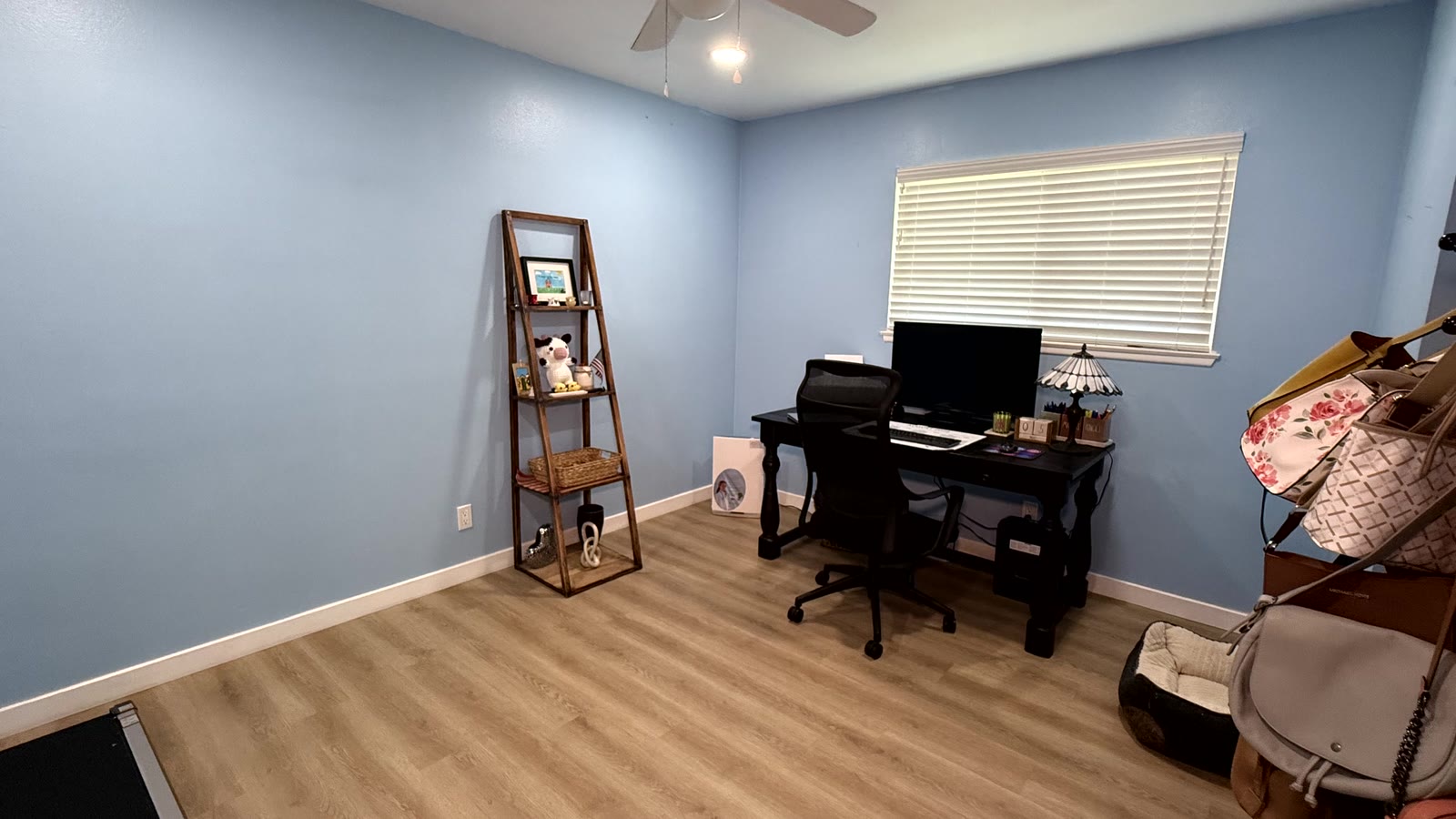 Third bedroom finished in light blue paint and currently used as a home office, with a ceiling fan, a window with white horizontal blinds and wood-look plank flooring.