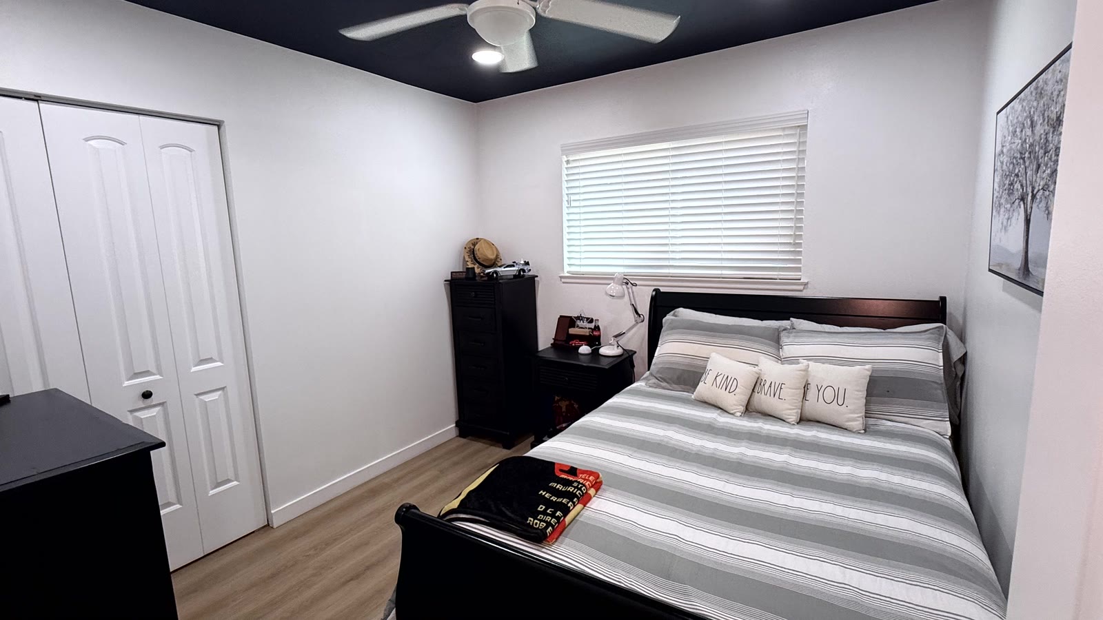 Second bedroom with white walls, a dark navy painted ceiling, a white ceiling fan, white bi-fold closet doors and wood-look plank flooring.