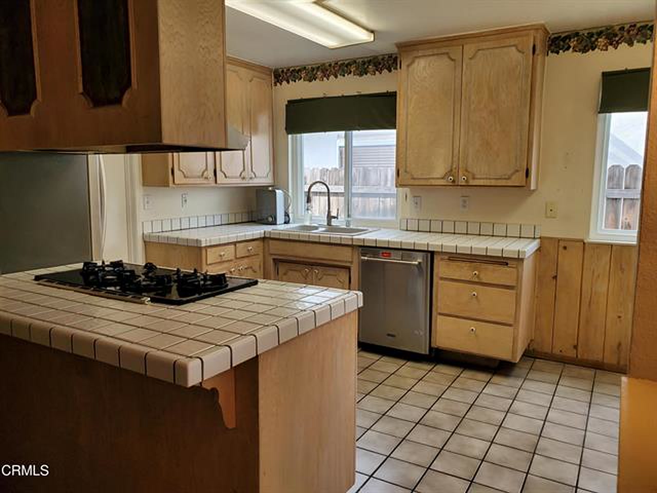 The kitchen in 2022: light oak cabinetry, tile counters with a bullnose edge, a peninsula cooktop with an overhead hood, and tile flooring.