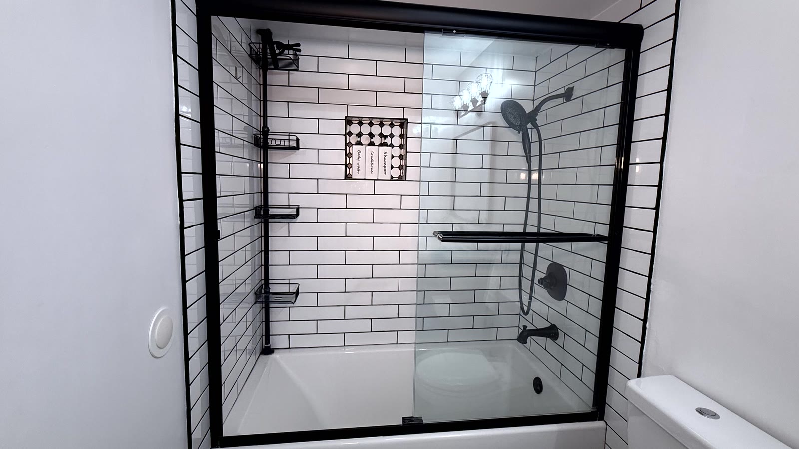 Hall bath tub and shower combination with black-framed sliding glass, white subway tile with dark grout to full height, a mosaic-backed recessed niche and black fittings.