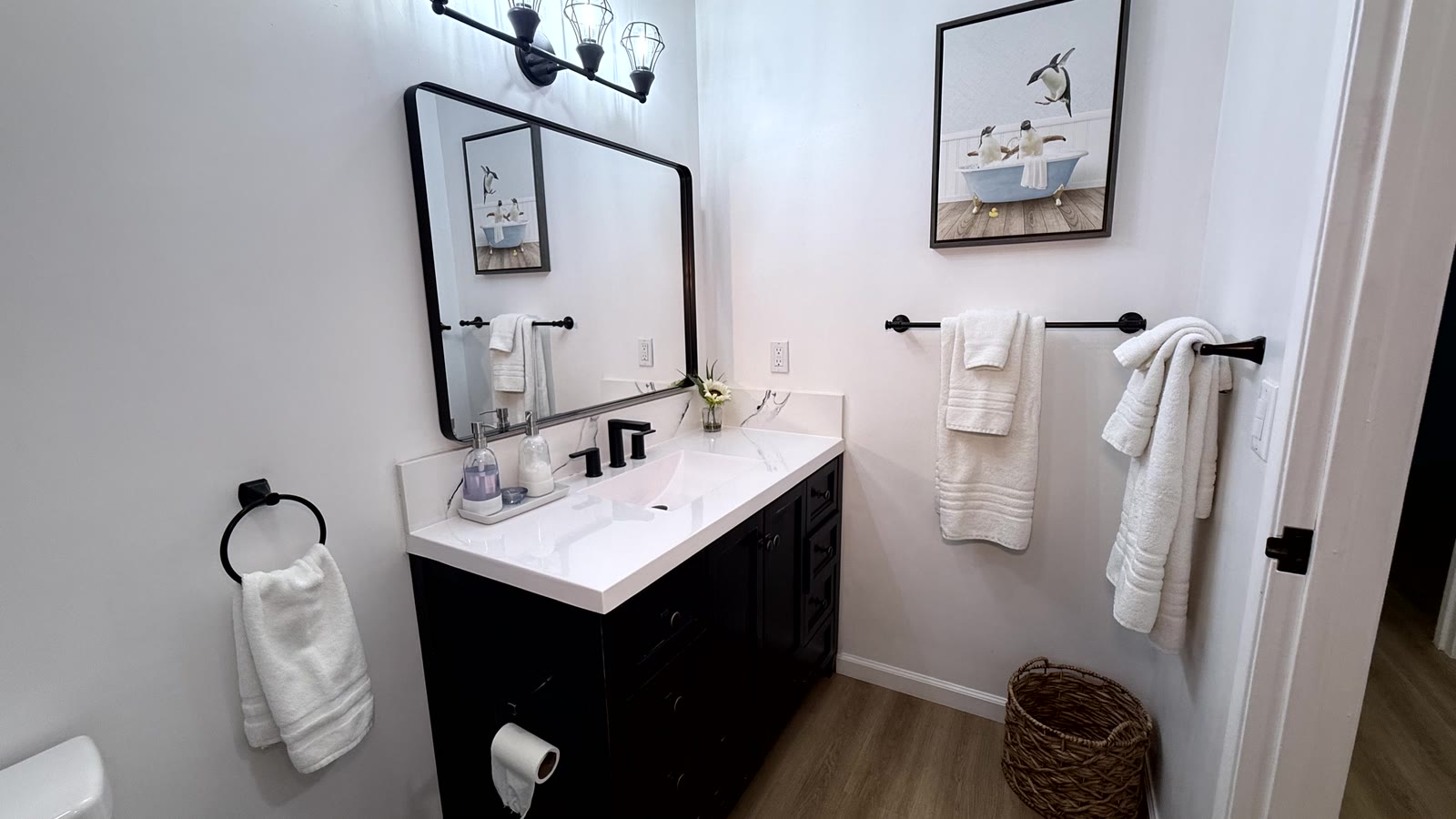 Hall bath single-sink vanity: dark raised-panel cabinet, white stone-look top, undermount rectangular sink, black widespread faucet, rounded-rectangle black-framed mirror and a four-light black caged vanity bar.