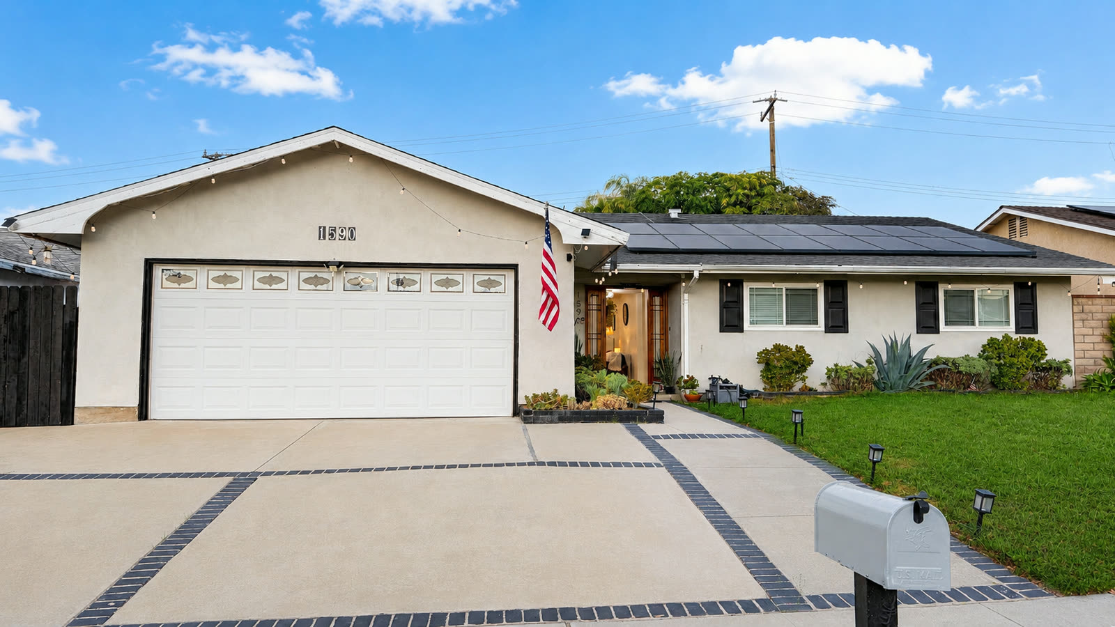 1590 Sabina Circle seen from the driveway: a single story stucco ranch with a gabled roof, white sectional garage door, recessed entry with a wood door, black shutters, lawn and succulent beds.