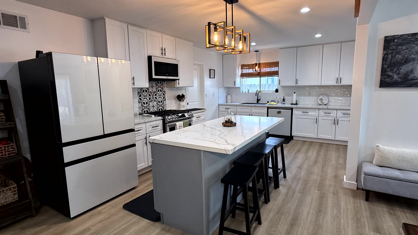 Kitchen with a center island under a black-and-brass linear pendant, thick waterfall-edge stone-look counter, black counter stools, white shaker cabinetry stacked to the ceiling and a stainless gas range.