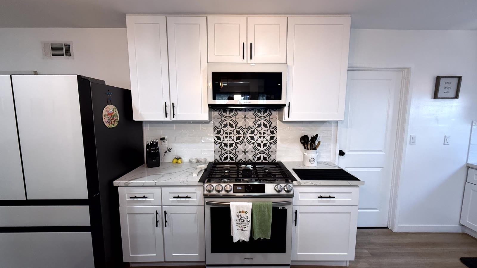Range wall square on: stainless five-burner gas range with an over-range microwave, a panel of patterned encaustic-look tile behind it, stone-look counters and white shaker cabinets with black bar pulls.