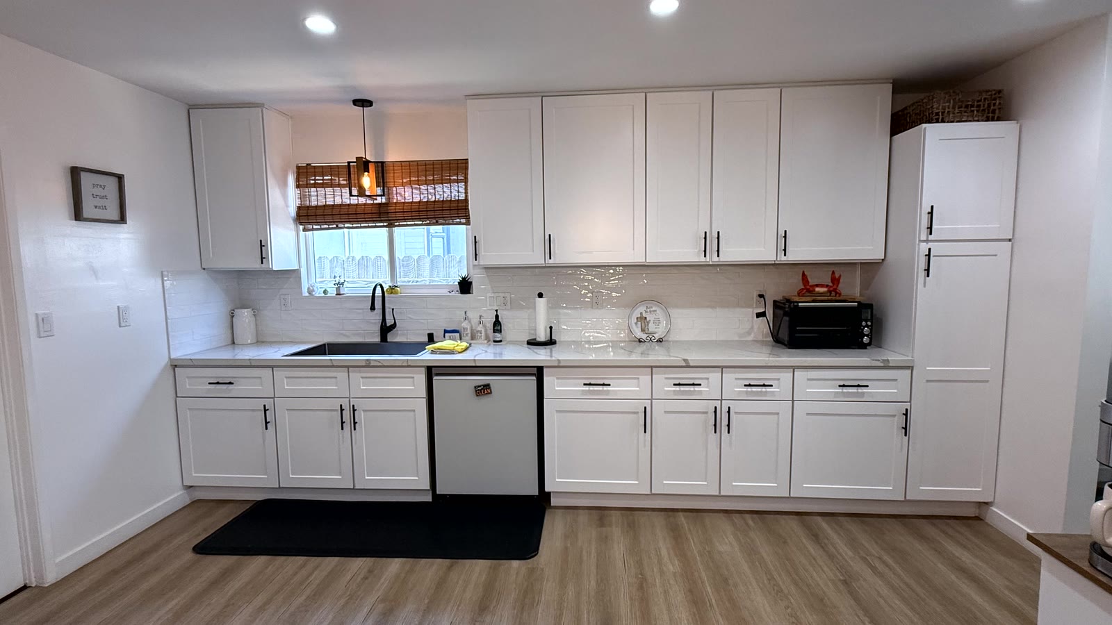 Kitchen sink run: white shaker base and wall cabinets with black bar pulls, tall pantry cabinet, stone-look counters, glossy white stacked-tile backsplash and a black undermount sink under a window.