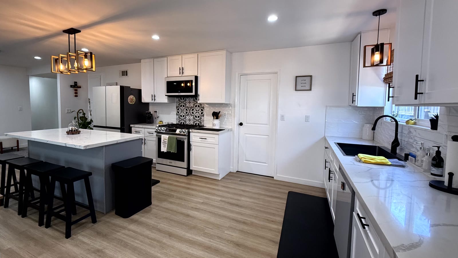Kitchen seen wide from the dining side: island with counter stools at left, stainless gas range and patterned tile at center, sink run at right and pendant lighting above.