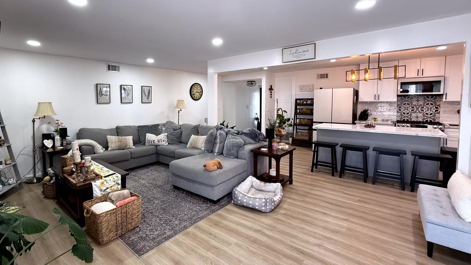 Wide view of the living room open to the kitchen across a broad cased opening, with the island, counter stools and linear pendant beyond, and continuous wood-look plank flooring throughout.