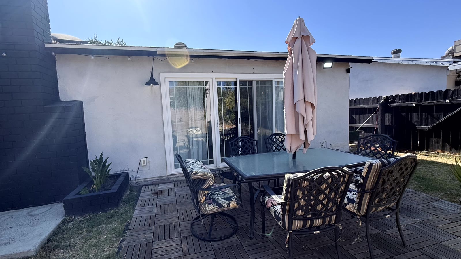 Rear patio off the dining room slider: interlocking wood deck tiles, a cast-aluminum dining set with six cushioned chairs and a closed umbrella, string lights on the eave and a black barn-style wall sconce.