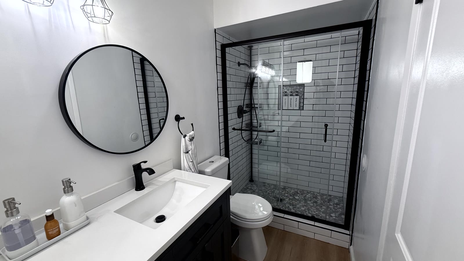 Primary bath walk-in shower with black-framed sliding clear glass, white subway tile with dark grout, a pebble mosaic floor, recessed niche and a black rain head.