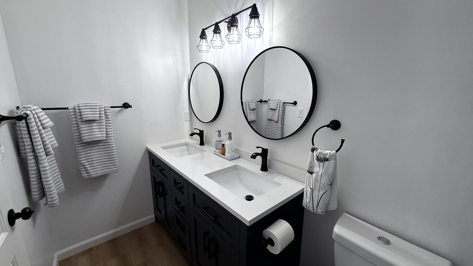 Primary bath double-sink vanity: dark cabinet with fretwork panel detail, white stone-look top, two undermount sinks with black faucets, two round black-framed mirrors and a four-light black caged vanity bar.