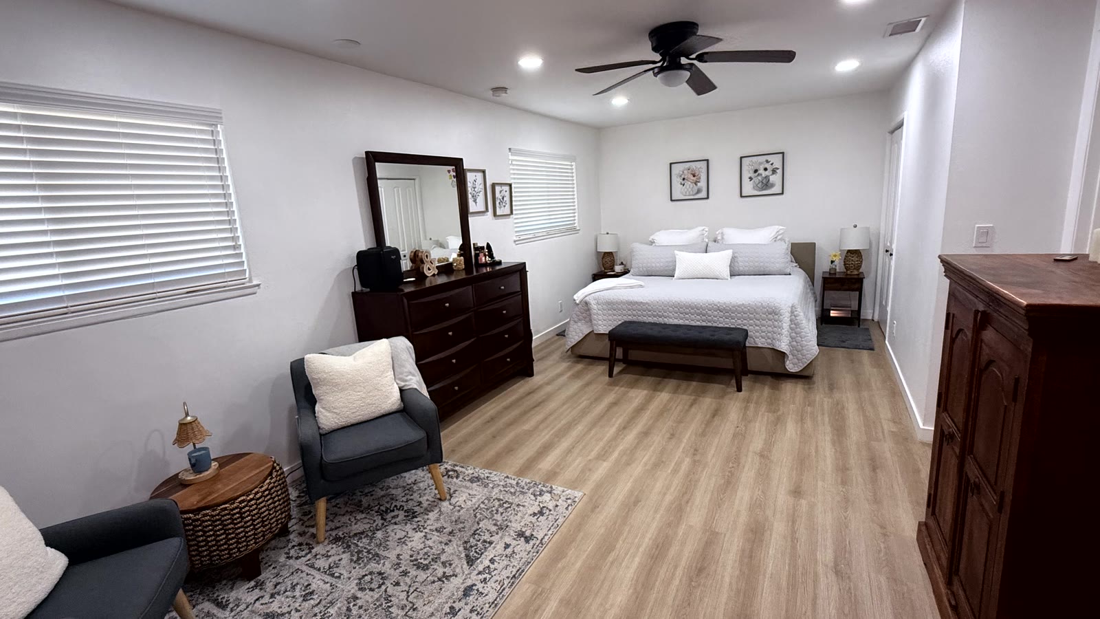 Primary bedroom with a ceiling fan, recessed lighting, two windows with white horizontal blinds, white walls and wood-look plank flooring.